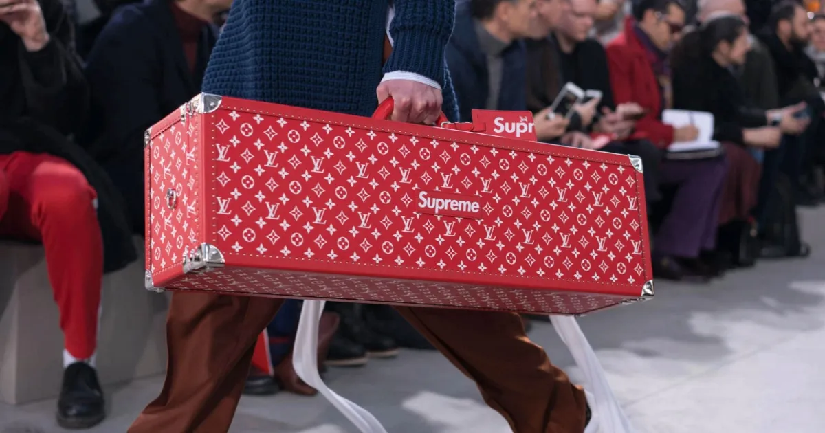 A man carrying the luxury and streetwear collaboration between Supreme and Louis Vuitton red trunk with the brands' logos in white
