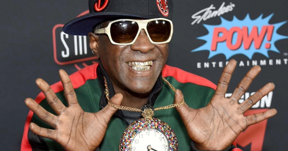 Black rapper Flava Flav wearing teeth jewelry, big sunglasses, and a biggold chain