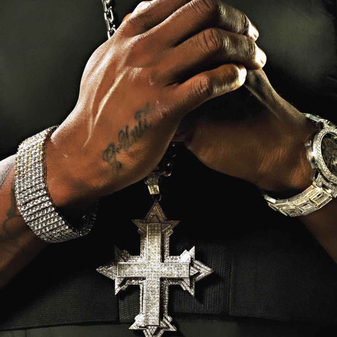 A black man wearing a black shirt with silver jewelry, a crucifix chain on the neck representing Hip-Hop and Religion